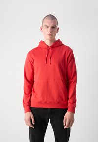 Felpa rossa con cappuccio realizzata in tessuto morbido, presenta una tasca frontale, cappuccio con coulisse e un piccolo logo sul petto. Abbinata a pantaloni neri.