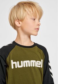 Grøn og sort langærmet skjorte med hvidt "hummel"-logo, der har raglan-design og kontrasterende skulderstriber. Glat stoftype.