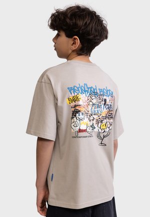 Dreng med krøllet hår iført beige T-shirt med farverig graffiti-stil kunst og tekst "I’m okay," "smile," og "contemporary streetwear" på ryggen.