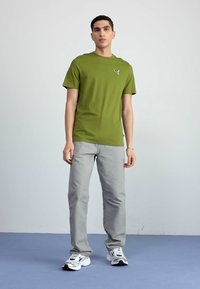 Olivengrønn t-shirt med korte ærmer og en lille hvid logo, parret med lysegrå lige ben bukser og hvide sneakers. Enkel design.