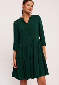 Kobieta w ciemnozielonej, warstwowej sukience do kolan z rękawami 3/4 i złotymi kolczykami w kształcie kół, na jednolitym jasnym tle.