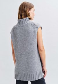 Grauer gestrickter ärmelloser Pullover mit hohem Kragen, geripptem Saum und seitlichen Schlitzen, der eine strukturierte Oberfläche und eine lockere Passform zeigt.