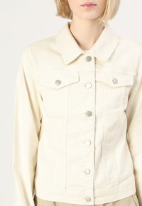 Veste en denim beige clair avec un col, des poches avant et des boutons-pression en métal argenté. Texture lisse et coupe ajustée, se terminant à la taille.