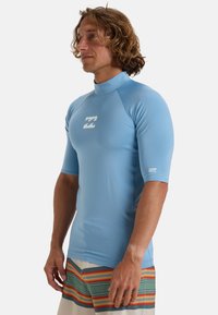 Blå kortærmet rash guard lavet af strækbart materiale med høj hals og hvidt logo på brystet. Har en glat, let tekstur.