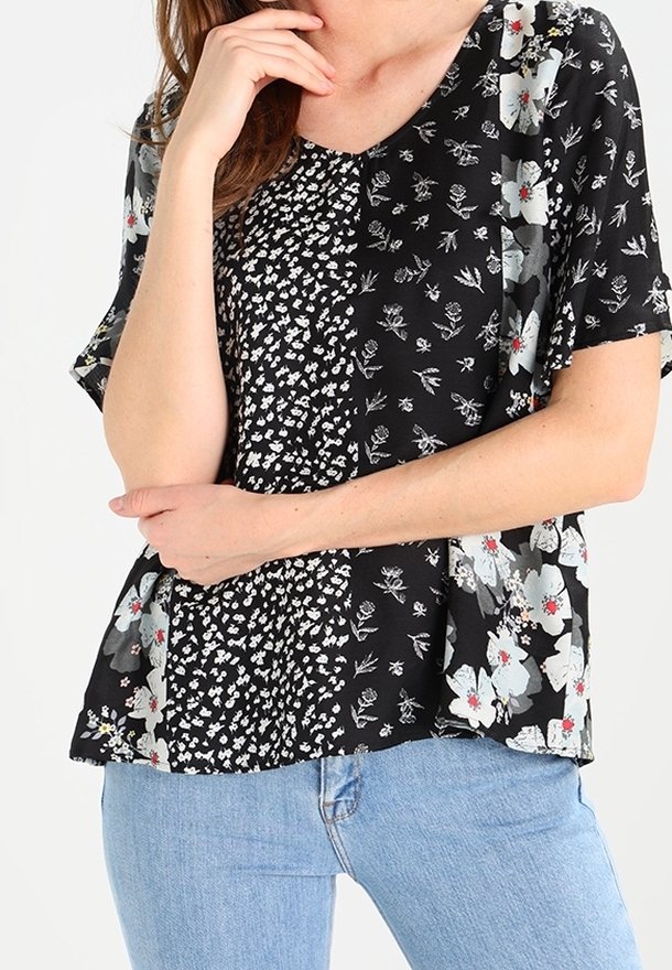 Femme portant un chemisier noir à manches courtes avec des motifs floraux blancs mélangés et un jean bleu clair, main reposant sur le menton et bras croisés.
