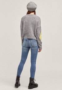 Grijze gebreide trui met gele accenten, blauwe skinny jeans en zwarte veterlaarzen. De trui heeft een boxy pasvorm en een geribde zoom.