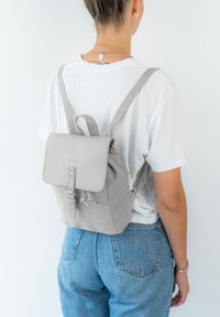 Harmaa mokkareppu, jossa on jämäkkä läppä, säädettävät hihnat ja etupuolen solki. Minimalistinen muotoilu ilman näkyviä kuvioita tai metalliosia.