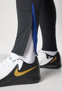 Pantaloni atletici neri a coste con dettaglio della cerniera blu abbinati a scarpe Nike bianco e nere, con il classico swoosh dorato e design testurizzato.