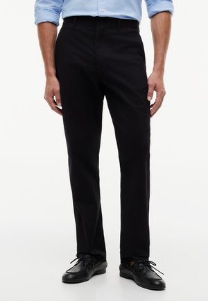 Homme portant un pantalon noir droit, des chaussures noires à lacets et une chemise bleu clair à manches longues avec les manches retroussées.