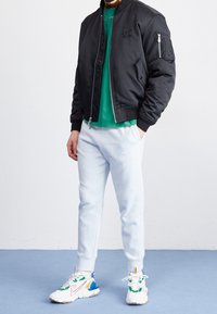 Svart bomberjacka med dragkedja, grön T-shirt under, ljusblå sweatpants och mångfärgade sneakers med olika accenter och texturer.