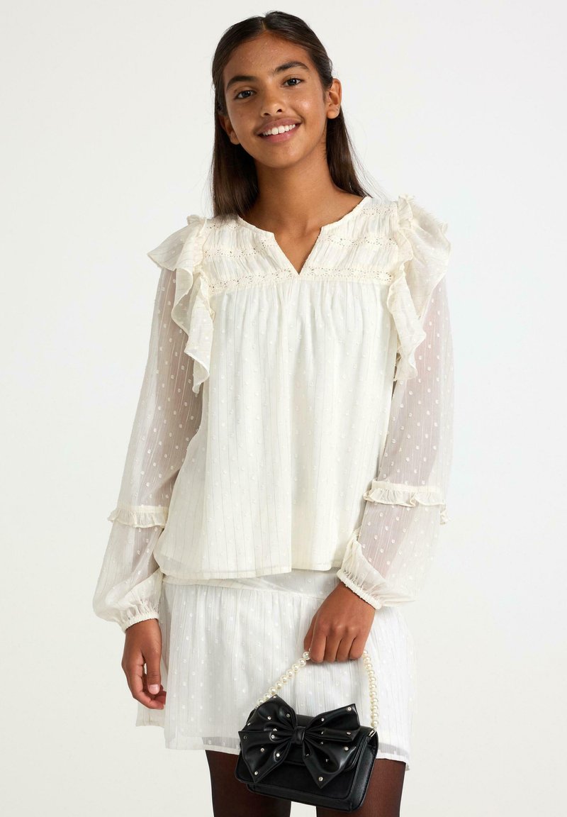 Ung kvinde i gennemsigtig hvid bluse med volanger og nederdel, der holder en sort pung med perlehank og stor sløjfe, mens hun smiler mod en hvid baggrund.