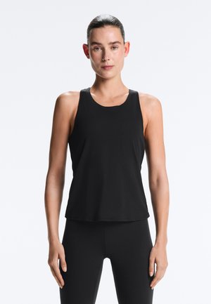 Kvinde med krepperet hår tilbage, iført en ærmeløs sort tanktop og sorte leggings, stående mod en ensfarvet lys baggrund.