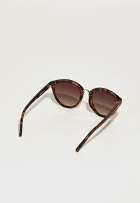 Ochelari de soare cu un montaj țesut din tortoise, cu o formă de ochi de pisică, lentile maronii cu un gradient și brațe subțiri cu accente de hardware aurii discrete.