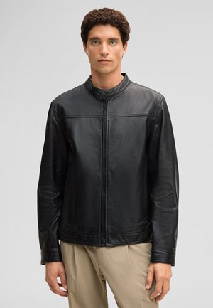 Mann steht, nach vorne blickend, trägt eine schwarze Lederjacke mit Reißverschluss und Stehkragen sowie beigefarbene Hose vor einem schlichten hellgrauen Hintergrund.