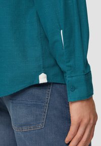 Teal geweven shirt met een knoopmanchet en zijslit, met een contrasterende witte accent aan de zoom. Denim jeans zichtbaar erachter.