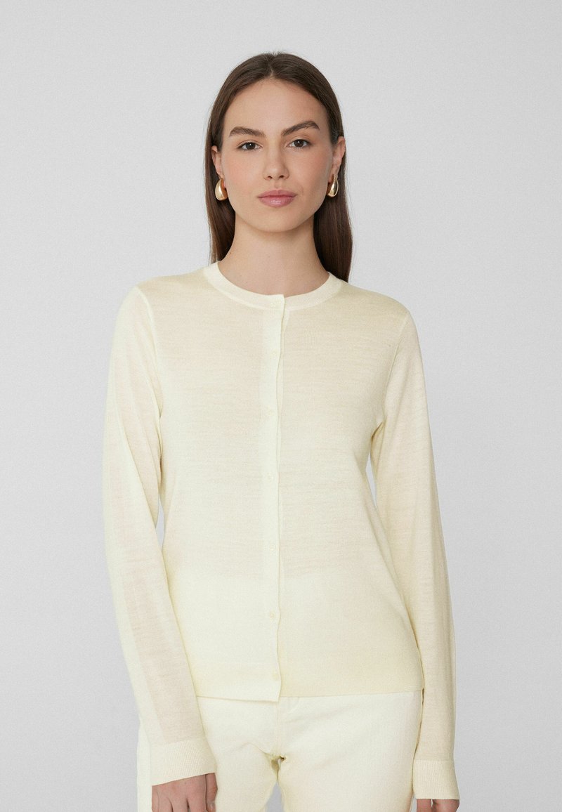 Cárdigan tejido de color amarillo claro con mangas largas, cuello redondo y cierre frontal con botones. Textura suave con puños y dobladillo acanalados.