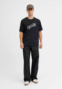 Must küllane t-särk valge disainiga, millele on kombineeritud tumedad teksapüksid ja valged tossud. Mudel kannab halli mütsi ja seisab heleda tausta ees.