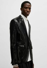 Schwarzer Paillettenblazer mit einem eleganten Design, featuring einen Einzelknopfverschluss, Reverskragen und zwei Fronttaschen. Getragen über einem weißen Hemd.