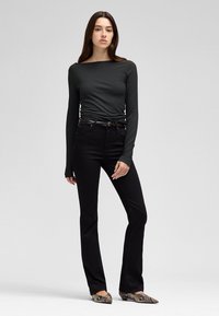 Vrouw staand met lang donker haar, draagt een aansluitende donkergrijze top met lange mouwen, zwarte broek, zwarte riem en schoenen met slangenhuidmotief.