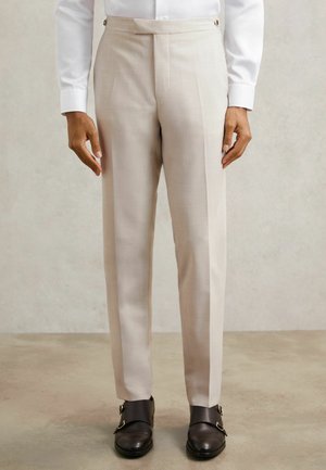 Beige op maat gemaakte broek met scherpe vouwen, gestructureerde stof en knopen aan de zijkant. Gecombineerd met donkerbruine schoenen met dubbele monksluiting.