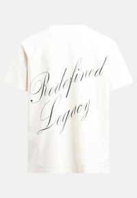 T-shirt en coton crème avec le texte "Héritage Redéfini" en lettres cursives noires au dos. Manches courtes et coupe décontractée.