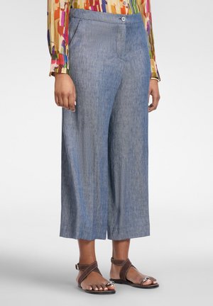 Lichtblauwe cropped broek met wijde pijpen, gedragen met bruine sandalen met enkelbandje en een kleurrijke blouse met lange, geplaatste mouwen en patroon.