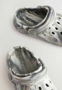 Sabots gris et blancs avec un motif, fabriqués en matériau synthétique ; dotés de perforations pour la ventilation et d'une sangle arrière pour un maintien sécurisé.