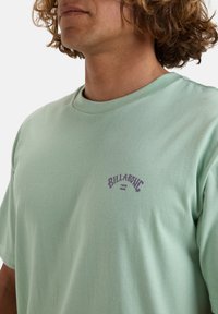 Muntgroene katoenen t-shirt met een ronde halslijn en een paarse "Billabong"-logo op de borst. Zachte textuur, comfortabele pasvorm.