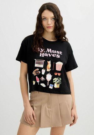Joven mujer con cabello castaño ondulado lleva una camiseta corta negra con la frase "My Must Haves" con iconos de colores y una minifalda plisada beige.