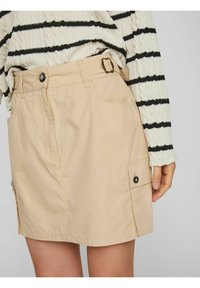 Beige nyttokjol med spännen, sidofickor och en något A-linjeformad silhuett. Tyget verkar mjukt med minimal textur.