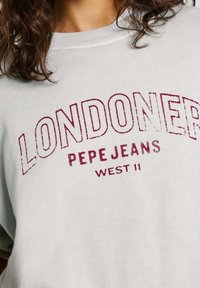 Sudadera gris claro con texto impreso en rojo que dice "LONDONER PEPE JEANS WEST 11." Presenta un cuello redondo y una textura suave.
