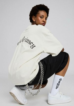 Persona joven con cabello corto y rizado agachada, vistiendo una camisa blanca de gran tamaño, pantalones cortos negros, calcetas blancas con texto, accesorios de cadenas y zapatillas blancas.