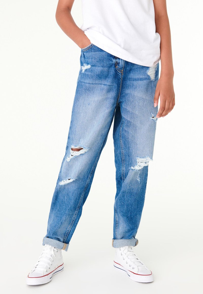 Person wearing light blue ripped jeans with rolled cuffs, white sneakers, and a white short-sleeve shirt, standing with one hand in pocket.