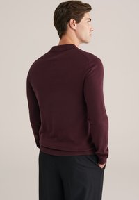 Férfi oldalra nézve, sötét burgundi színű, testhez simuló, hosszú ujjú pulóverben és fekete nadrágban, kezével a zsebében, semleges háttér előtt.