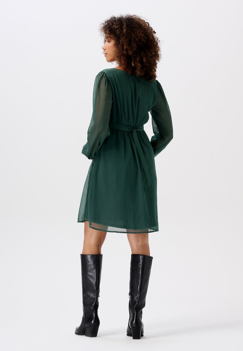 Grünes, halbtransparentes Kleid mit langen, gepufften Ärmeln und tailliertem Bund, kombiniert mit knielangen schwarzen Lederstiefeln.