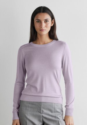 Kvinde med langt mørkt hår iført en lys lilla langærmet sweater og grå bukser, stående mod en ensfarvet hvid baggrund.