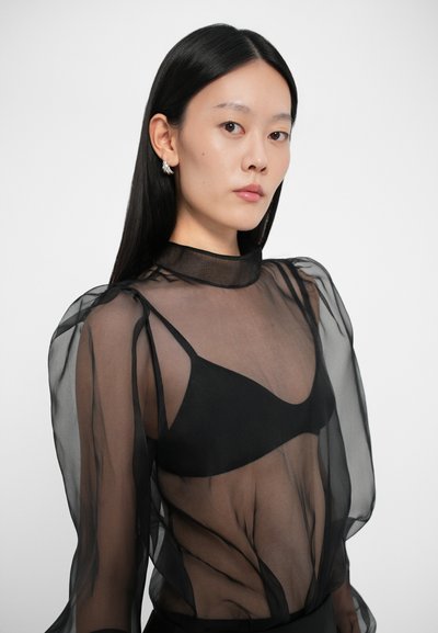 Musta läpinäkyvä paita puffihihnoilla, korkea kaula-aukko ja tiukka musta bralette alla. Sujuvaa, kevyttä kangasta, jolla on läpikuultava laatu.