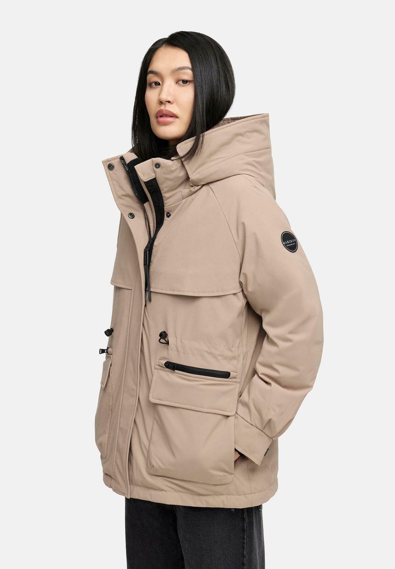 Beige insulated jacket with a high collar, large front pockets, and a zip closure. Features black hardware and a logo patch on the sleeve.