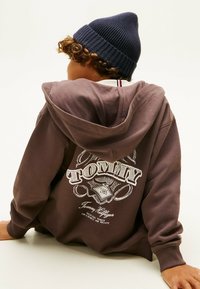 Sudadera marrón con un gran logo gráfico blanco "TOMMY" en la parte posterior, con un cuello y puños de canalé. Combinada con un gorro azul marino.