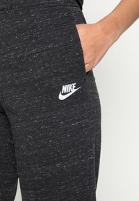 Hand i fickan, klädd i mörkgrå brokiga Nike-mjukisbyxor med vitt logo på låret, mot en neutral bakgrund.