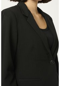 Blazer nero su misura con colletto strutturato, chiusura a un bottone, tasche a toppa e texture liscia, abbinato a un top nero sottostante.