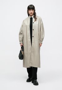 Žena v béžovom dlhom kabáte cez čierne nohavice a topánky, drží malú čiernu kabelku na jednoduchom svetlom pozadí.