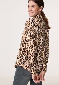 Bluse med leopardmønster i let vævning og lange ærmer, med sorte pletter på en lys brun bund, glat tekstur og knappedetaljer ved ærmerne.