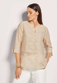 Beige-Tunika mit einem strukturierten Muster aus vertikalen Streifen, V-Ausschnitt und Dreiviertelärmeln. Kombiniert mit weißen Hosen.