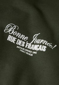 Mørkegrønt stof med hvid trykt tekst: "Bonne Journée! RUE DES FRANÇAIS," etableret 1992, med en glat tekstur.