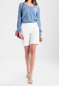 Donna che cammina indossando una blusa blu a maniche lunghe a fantasia, pantaloni corti bianchi fino al ginocchio, tacchi beige con cinturini e porta una piccola borsa rossa.