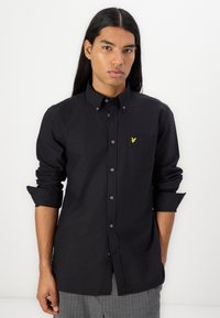 Camicia nera con bottoni realizzata in cotone, dotata di una tasca sul petto e un piccolo logo giallo. Maniche lunghe arrotolate leggermente ai polsini.