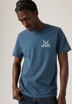 Jeune homme aux cheveux tressés regarde vers le bas, portant un t-shirt bleu Levi's avec un logo oiseau blanc sur la poitrine.