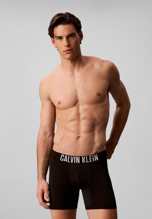 Man zonder overhemd met kort donker haar die zwarte Calvin Klein boxers draagt tegen een effen lichtgrijze achtergrond.