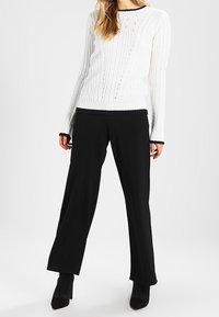 Biały sweter z warkoczowym splotem z granatowym obszyciem przy dekolcie i mankietach, zestawiony z czarnymi szerokimi spodniami i czarnymi botkami na obcasie.